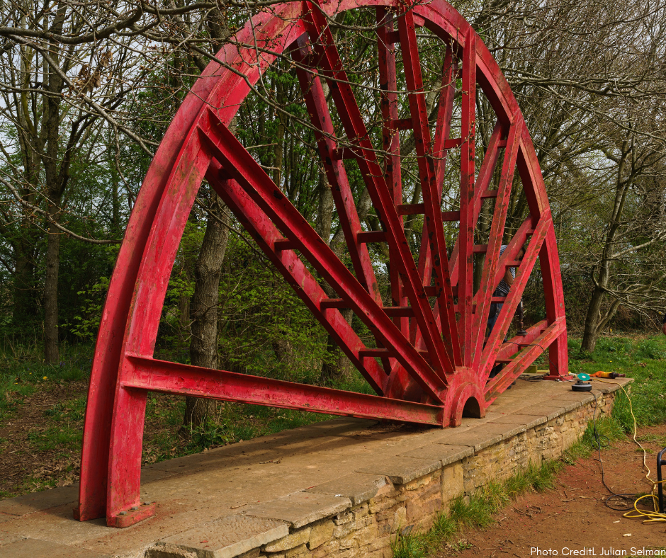 Photograph of the Centenary Wheel before its refurbishments. It is a large, red, semicircular metal wheel on a low stone wall. It is clearly rusted and weather-worn and dirty. 