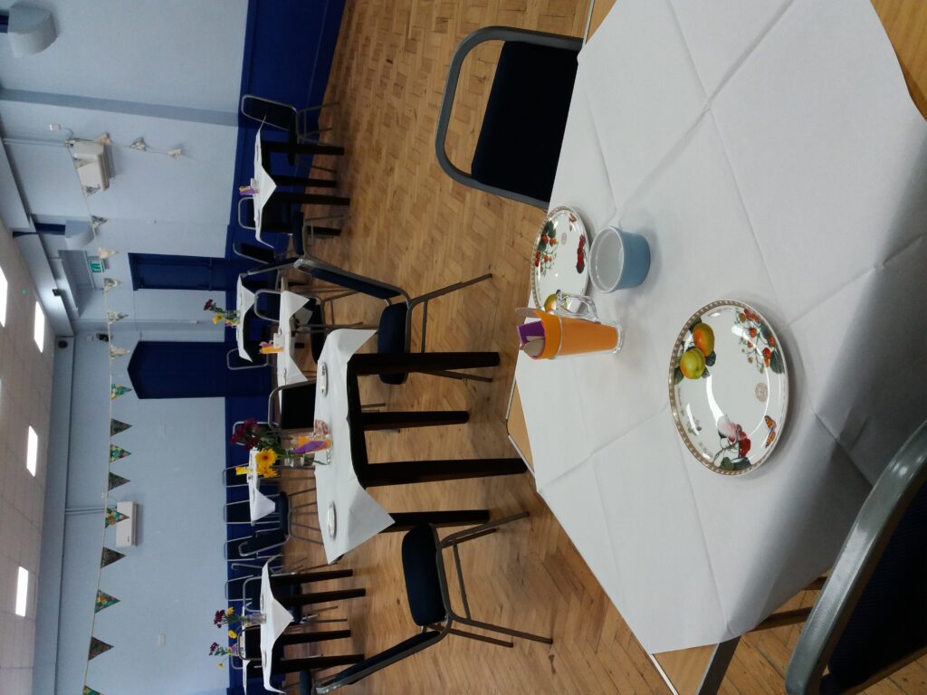photograph of an empty hall prepared for an afternoon tea. Bunting is strung up, there are several tables adorned with white tablecloths and fine china.