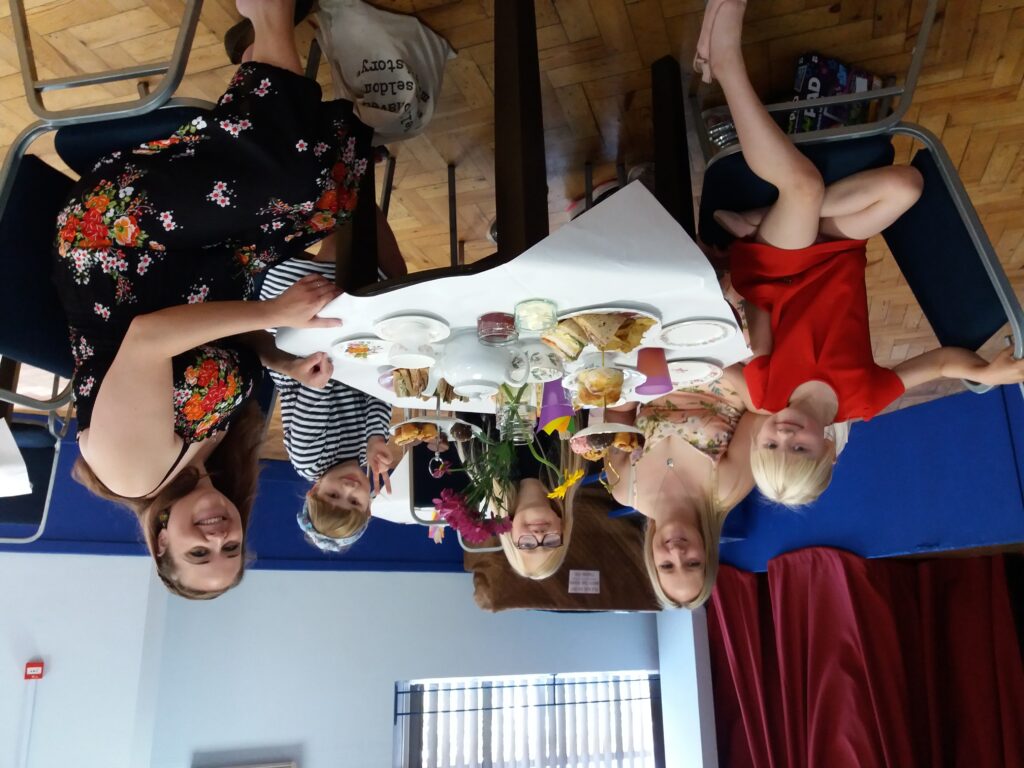 A photograph of a group of people sitting around a tea table. There are two adults and three children, they all face the camera, smiling. Their table has plates of cake, scones, and tea on it.