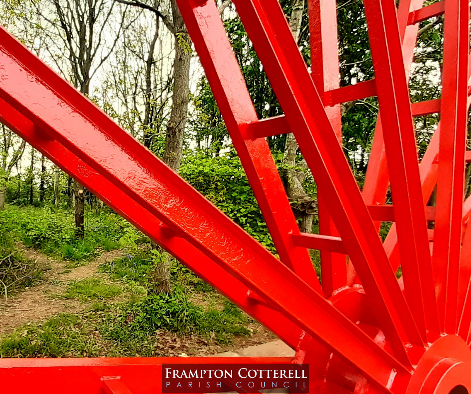Close up photograph of the spokes of the Centenary Wheel. They are shiny and red. Behind the spokes is a green wooded area.