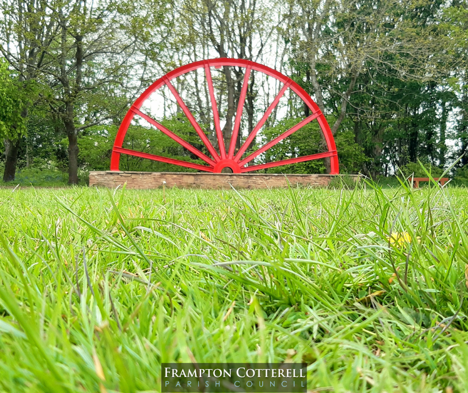 Photograph of the Centenary Wheel from a distance. The wheel is bright red against the dark green of trees behind and the bright green of grass in front.