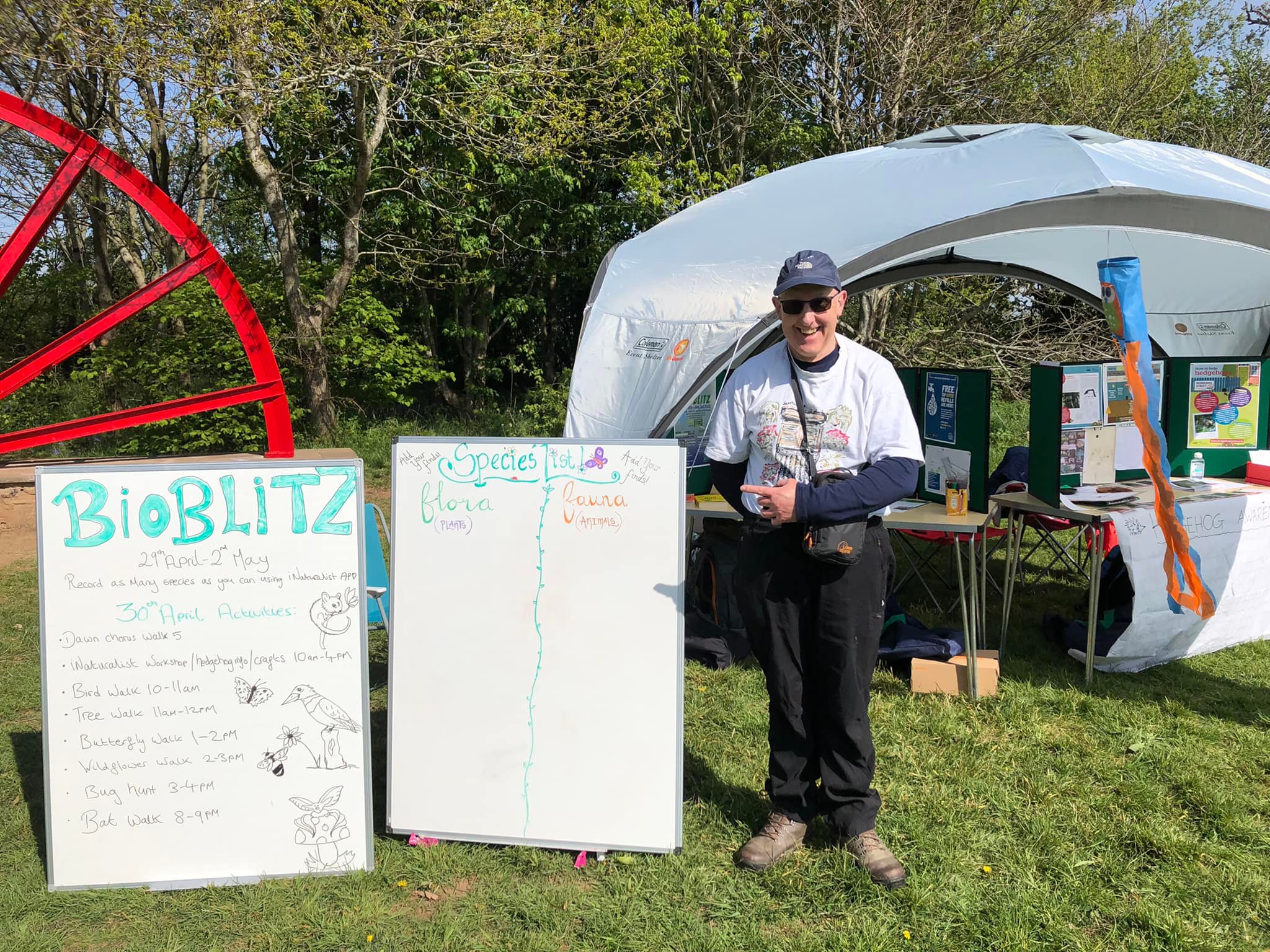 photograph of councillor Julian Selman standing in front of the Bioblitz stall, beside two white boards with Bioblitz activities listed on one, and SPECIES LIST Flora and Fauna written on the other. The day is sunny, and the Centenary wheel can be seen to the left edge. 