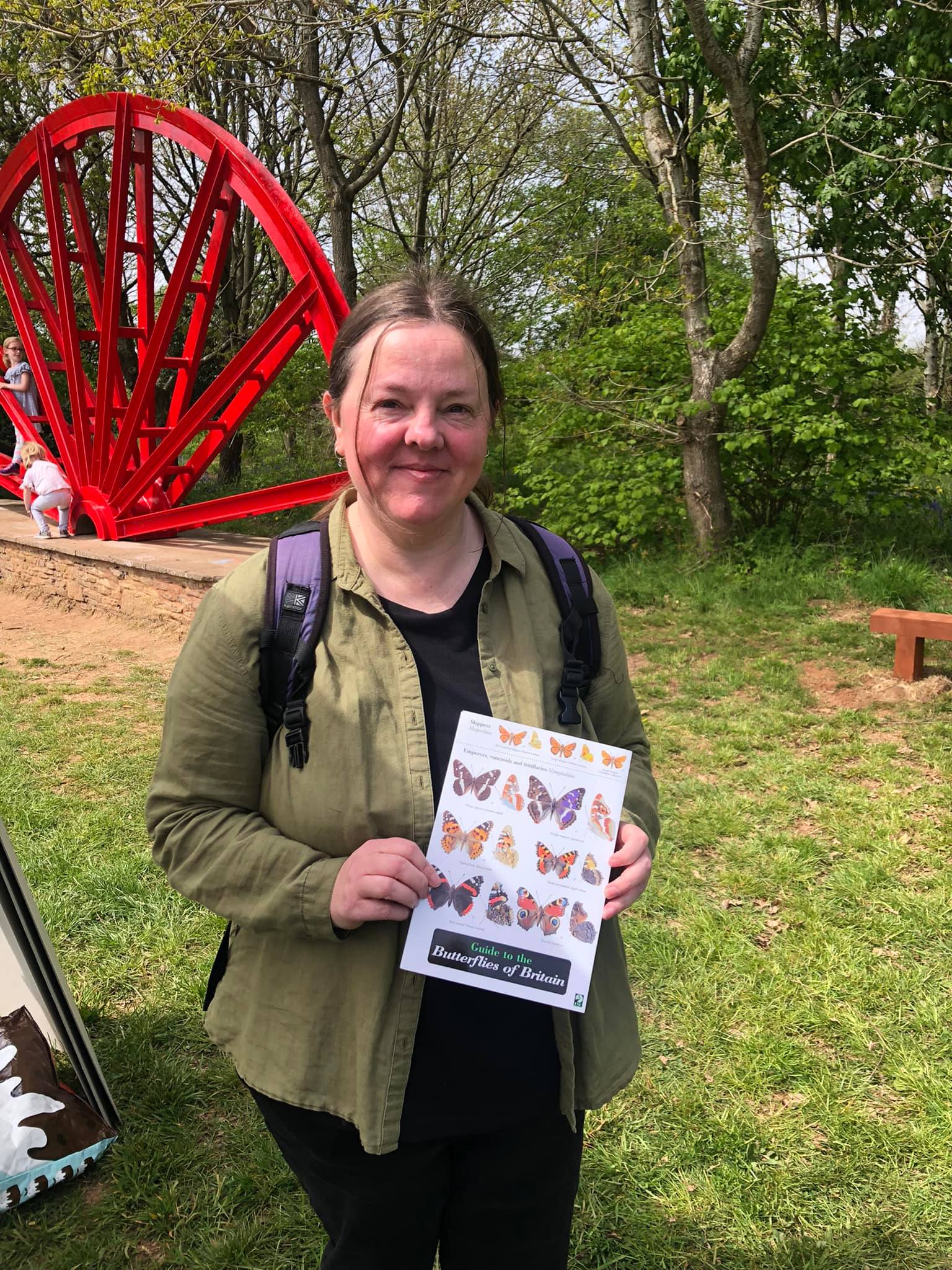 A person stands and smiles in front of the red Centenary wheel. They are holding a butterfly guide toward the camera.