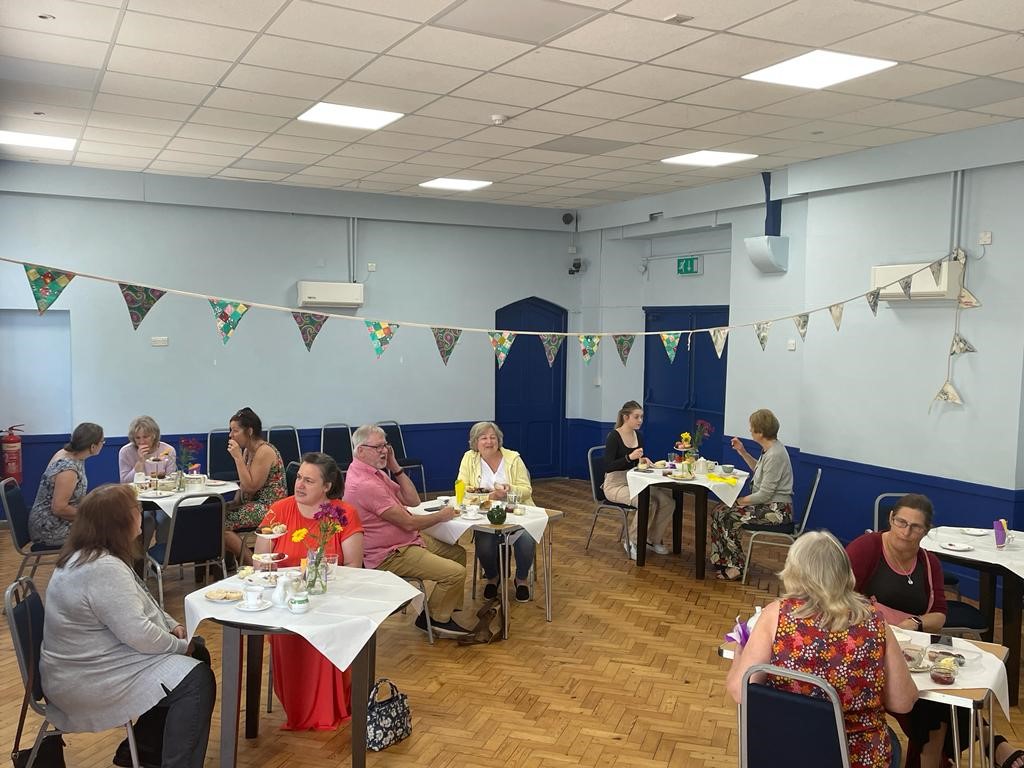 A photo of people sitting at tea tables in an indoor hall. There is bunting strung up above them, and they sit at tables with white table cloths. Each table also has tea and cakes on it.