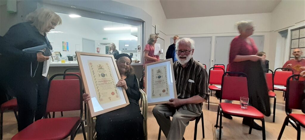  a photograph of Pat and Dave Hockey holding their Freedom of the Parish awards. They are an older man and woman, seated, smiling. The awards are large calligraphic documents on cream paper, inside large wooden frames.