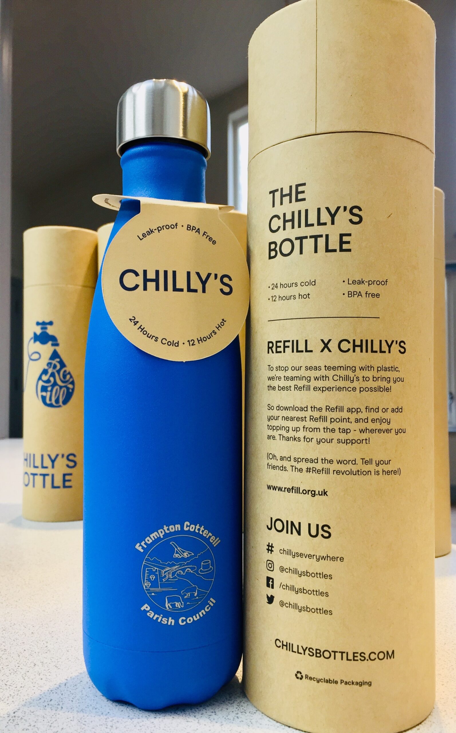 A photograph of a blue reusable water bottle, branded with "Frampton Cotterell Parish Council. It stands beside a "Chilly's" branded cardboard tube. On the tube. the following text is printed: The Chilly's Bottle. 24 hours cold. 12 hours hot. Leak Proof. BPA Free. Refill X Chillys. To stop our seas teeming with plastic, we're teaming with Chilly's to bring you the best Refill experience possible! So download the Refill app, find or add your nearest Refill point, and enjoy topping up from the tap - wherever you are. Thanks for your support! (Oh, and spread the word. Tell your friends. The #Refill Revolution is here!!) www.refill.org.uk JOIN US #ChillysEverywhere [Instagram logo] @chillysbottles [facebook logo] /chillysbottles [twitter logo] @chillysbottles chillysbottles.com
