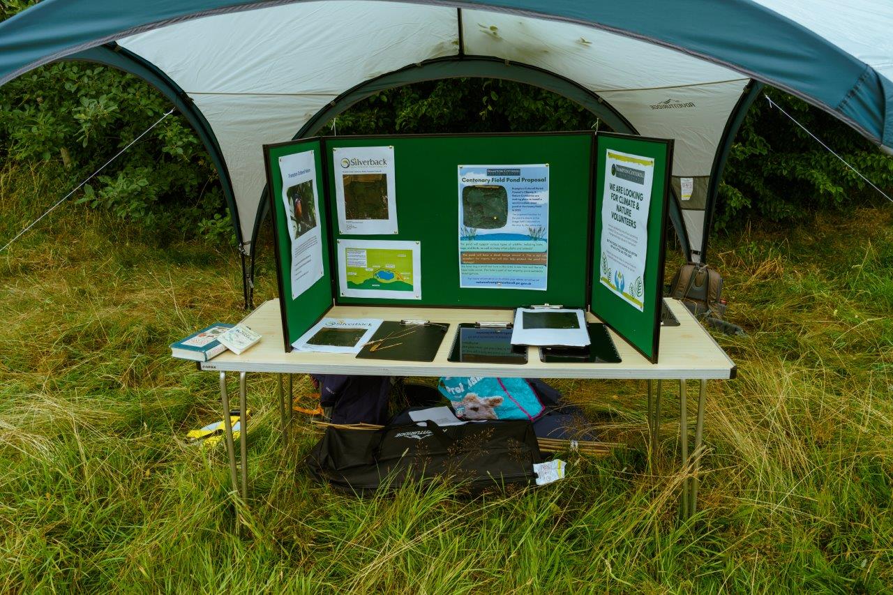 A pop up stall inside a gazebo in a field. There is a table with a large green three-panelled pin board with posters pinned to it.  