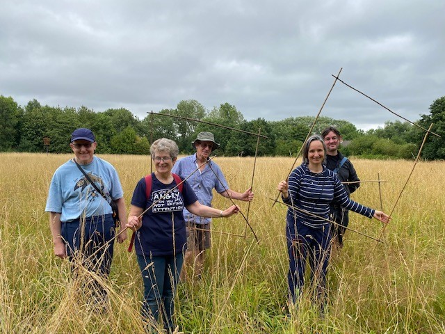 Photograph of five people standing in a hay meadow. They are holding squares made out of bamboo rods bound together at the corners.