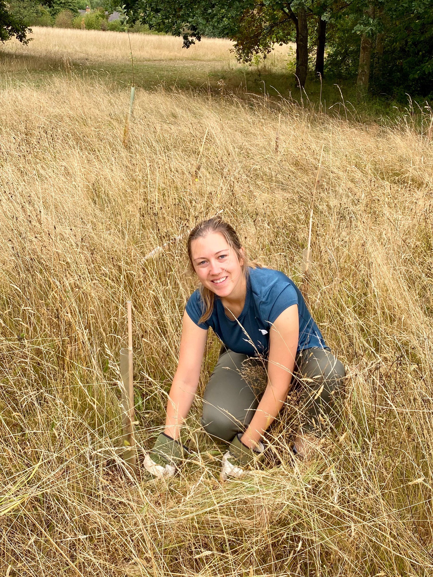 A young person with brown hair tied back in a ponytail smiles at the camera. They are crouching down in a field of tall, yellowing grass, beside a tree sapling. They are wearing a dark blue tshirt, khaki trousers, and green gardening gloves.