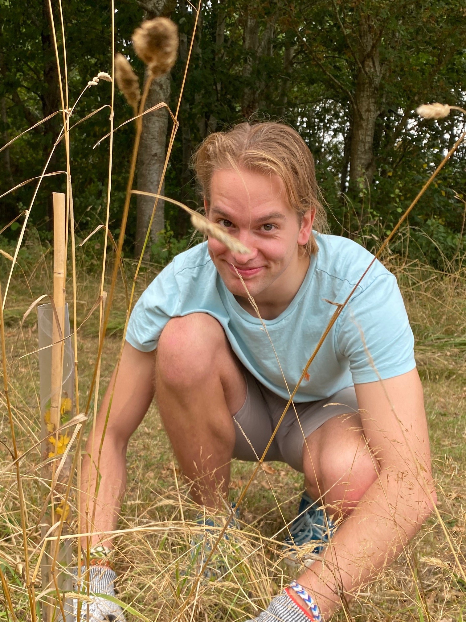 A young man with dark blonde hair pulled back in a ponytail, wearing a pale blue tshirt, khaki shorts, and grey gloves, crouches down in tall yellow-green grass beside a sapling tree. He is looking directly at the camera with a grin.
