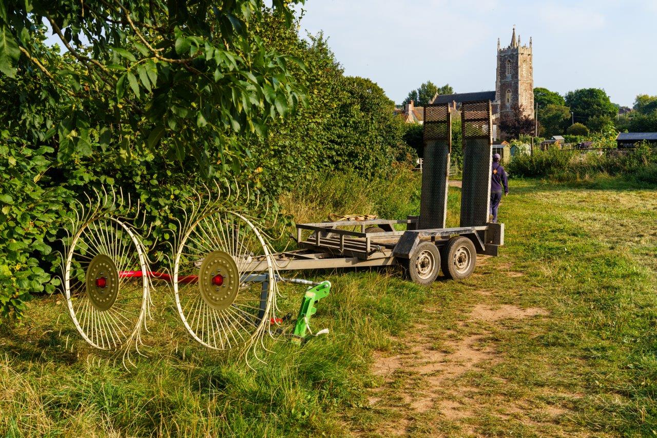 A field, a piece of machinery with large wheels ringed with some sort of curved fork, attached to what looks like a sort of flat bed trailer.