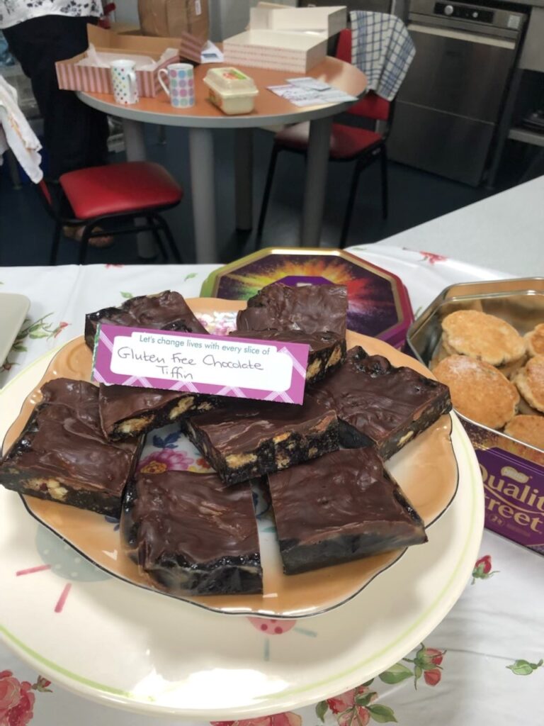 A plate of dark brown, rectangular brownies with biscuit pieces and purple swirls. The handwritten label reads, "Gluten Free Chocolate Tiffin".