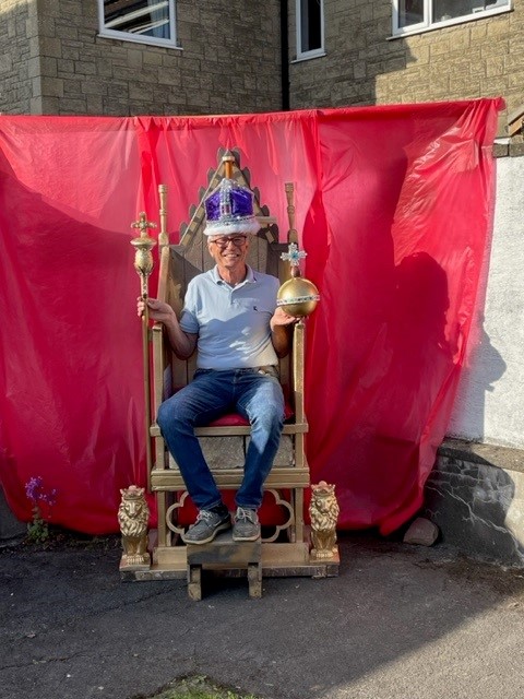 A man wearing a blue polo shirt and jeans sits in a golden throne. He has a purple crown on his head, and carries a royal orb in one hand and a royal sceptre in the other. He smiles broadly. The backdrop is a red cloth, in front of a house.