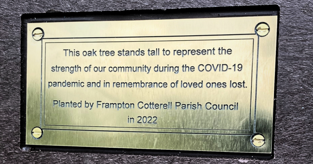 A gold plaque mounted on wood. The plaque reads, "This oak tree stands tall to represent the strength of our community during the COVID-19 pandemic and in remembrance of loved ones lost. Planted by Frampton Cotterell Parish Council in 2022."