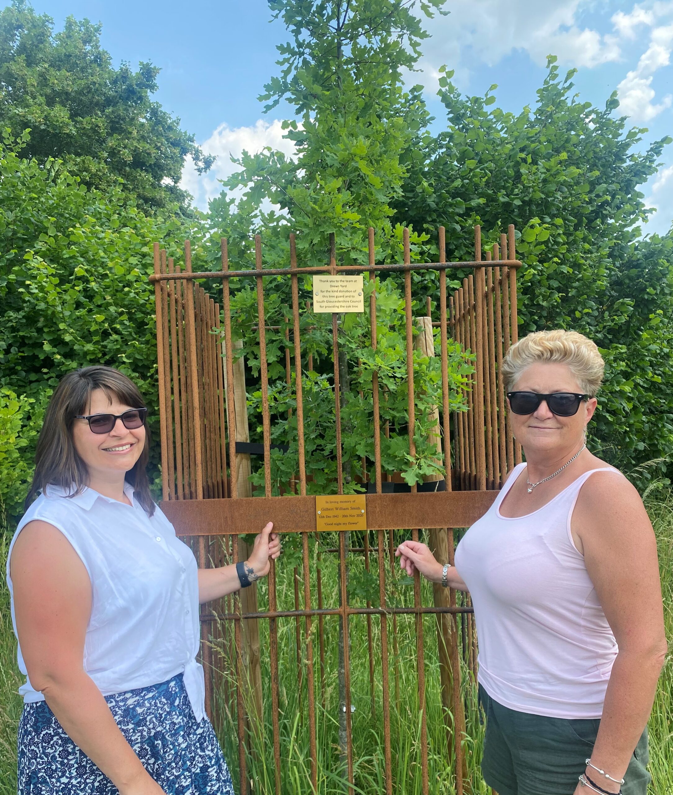 Two women, one with shoulder-length dark hair, one with short blonde hair, both wearing sunglasses, stand in front of a young oak tree. The oak tree is surrounded by bronze-coloured metal railings. These railings are about a foot higher than the women's heads. There are two small plaques attached to the railings. 