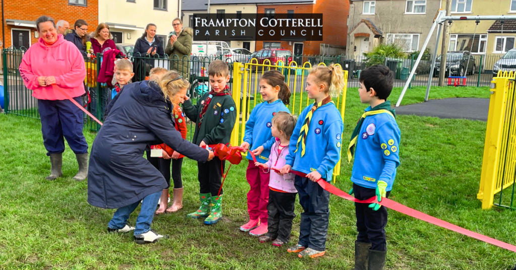 Frampton Cotterell Parish Council Chair, Councillor Linda Williams, cutting the ceremonial ribbon at Ridings Road Play Area. The ribbon is held up by a group of small children wearing Beavers uniforms. A group of people watches. Frampton Cotterell Parish Council logo.
