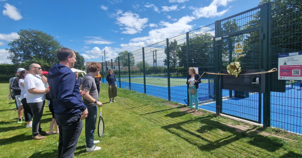 A crowd of people stand in the sunny Park to listen to Councillor Linda Williams give her speech to open the tennis courts.